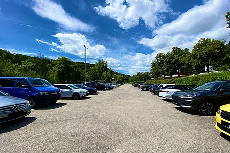 Parkplatz mit geparkten Autos auf beiden Seiten und bewaldetem Hintergrund unter blauem Himmel mit Wolken