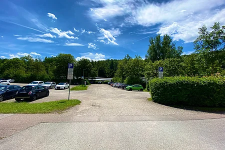 Parkplatz mit mehreren geparkten Autos, umgeben von Bäumen und blauem Himmel mit Wolken