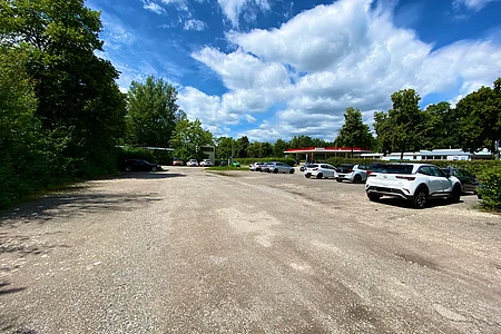 Großer Kiesparkplatz mit mehreren geparkten Autos und Tankstelle im Hintergrund unter blauem Himmel mit Wolken.