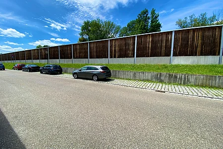Straße mit parkenden Autos, im Hintergrund Lärmschutzwand und Bäume unter blauem Himmel.