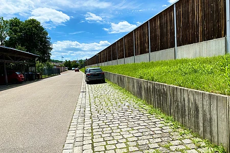 Straße mit gepflastertem Seitenstreifen, parkenden Autos, Holzsichtschutz und grünem Hang unter blauem Himmel.