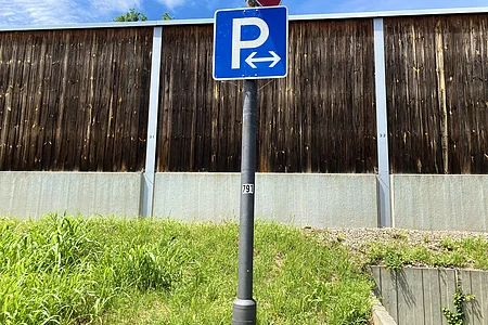 Blauer Parkplatzschild mit Pfeilen nach links und rechts an schwarzem Pfosten vor Holzwand und Grasfläche.