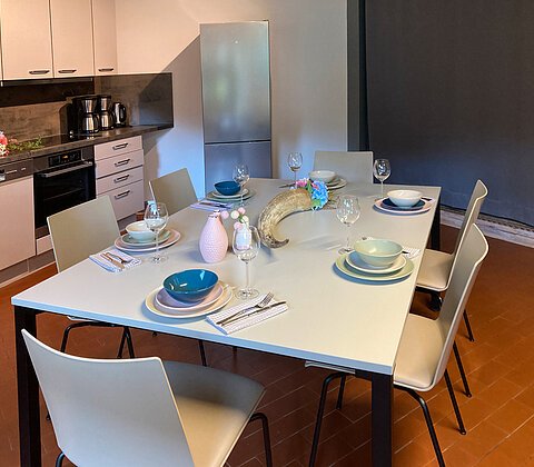 Esstisch mit sechs Stühlen, gedeckt mit Tellern, Gläsern und Besteck, in moderner Küche mit Kühlschrank und Herd.