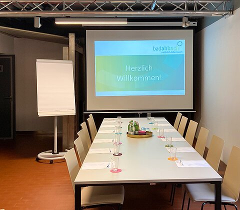 Konferenzraum mit langem Tisch, Stühlen, Flipchart und Projektionsleinwand mit Text „Herzlich Willkommen!“