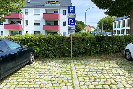 Parkplatz mit Kopfsteinpflaster, zwei Autos am Rand, Hinweisschilder für Parkregelungen vor Hecke und Wohnhäusern im Hintergrund