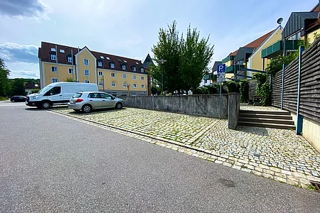 Parkplatz mit zwei Fahrzeugen vor gelben Wohnhäusern und Bäumen bei bewölktem Himmel