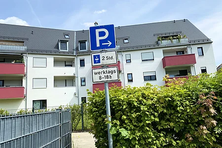 Parkplatzschild mit Pfeil nach rechts, zeitliche Begrenzung auf 2 Stunden werktags von 8 bis 18 Uhr vor Wohnhaus mit roten Balkonen