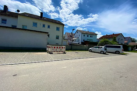 Parkplatz mit zwei geparkten Fahrzeugen und Absperrung vor Wohnhäusern unter blauem Himmel mit Wolken