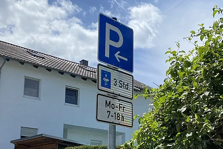 Verkehrsschild mit Parkhinweis links, Parkscheibe 3 Stunden, Mo-Fr 7-18 Uhr, Haus und Grünpflanzen im Hintergrund.