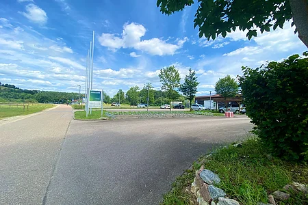 Parkplatz mit Autos und Fahrradständern bei blauem Himmel, links Wiese und Weg, rechts Bäume und Sträucher