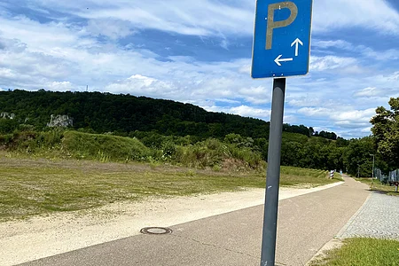 Blaues Parkzeichen mit Pfeilen nach links und geradeaus an einem Weg mit Wiesen und bewaldeten Hügeln im Hintergrund.