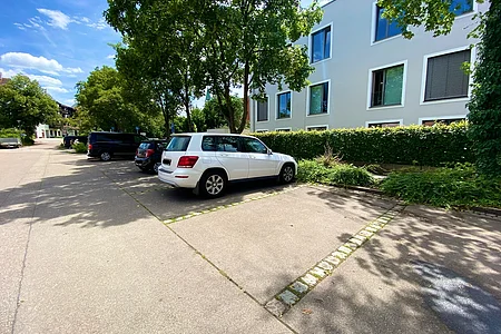 Parkplatz mit mehreren Autos neben einem weißen Wohngebäude und Bäumen bei sonnigem Himmel