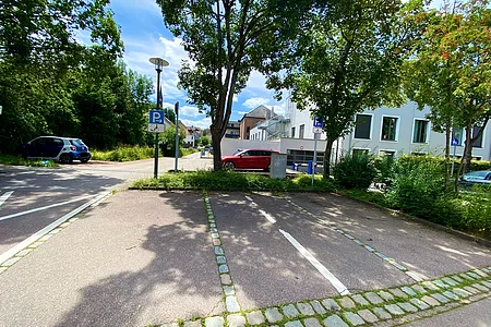 Leere Parkplätze mit Behindertenparkplatz-Schildern, Bäume und Gebäude im Hintergrund bei Tageslicht.