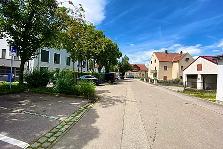 Straße mit geparkten Autos, Bäumen links und Wohnhäusern rechts unter blauem Himmel mit Wolken.