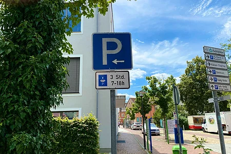 Parkplatzschild mit Pfeil nach links und Zusatzschild Parkscheibe 3 Stunden von 7 bis 18 Uhr an einem Gehweg vor Gebäuden und Bäumen
