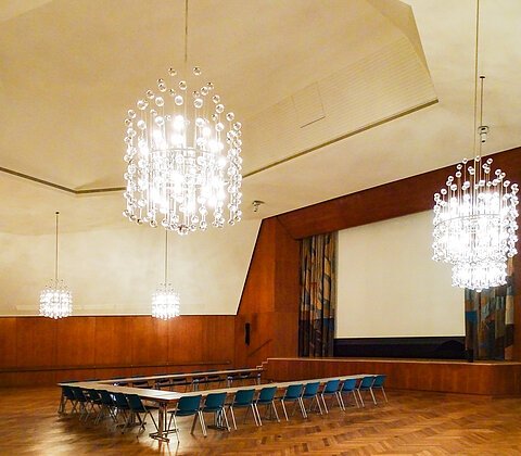 Großer Saal mit Holzvertäfelung, U-förmiger Tischanordnung und mehreren großen Kronleuchtern an der Decke.