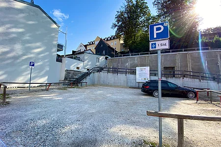 Parkplatz mit Schotterboden, zwei Parkschildern und einem schwarzen Auto, umgeben von Gebäuden und Bäumen