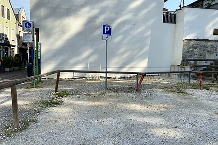 Parkplatz für Menschen mit Behinderung vor weißer Hauswand mit Absperrung aus Holzpfosten und Metallstange