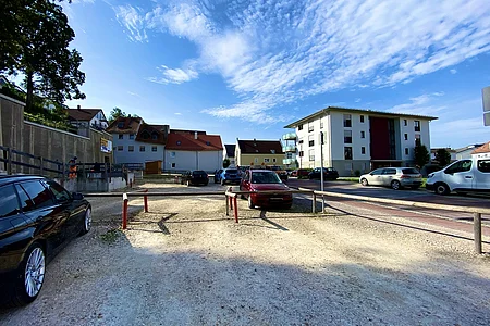 Parkplatz mit mehreren geparkten Autos vor Wohnhäusern unter blauem Himmel mit Wolken