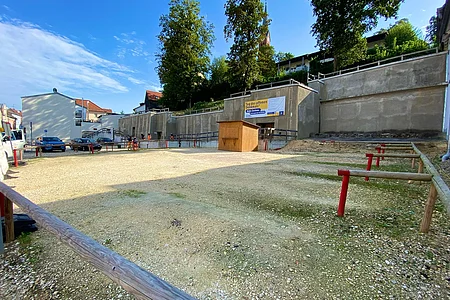 Freier, eingezäunter Platz mit Kiesboden vor einer Betonmauer und einem Holzhäuschen unter blauem Himmel.