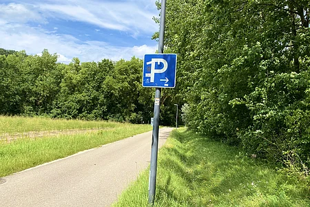 Blauer Parkplatzwegweiser an einer Straße mit Wiesen und Bäumen unter blauem Himmel.