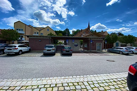 Parkplatz mit mehreren Autos vor einem kleinen Backsteingebäude und Häusern unter blauem Himmel mit Wolken