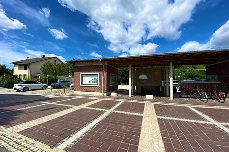 Parkplatz mit gepflastertem Boden, überdachtem Wartebereich, Fahrrädern und Autos unter blauem Himmel mit Wolken