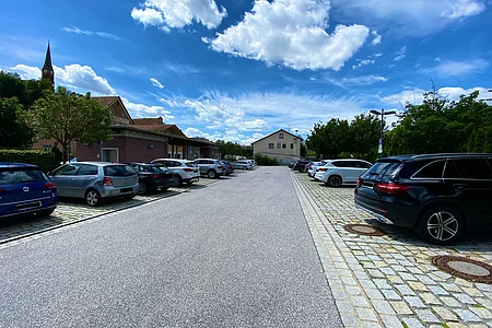 Parkplatz mit mehreren Autos, Bäumen und Gebäuden unter blauem Himmel mit Wolken.