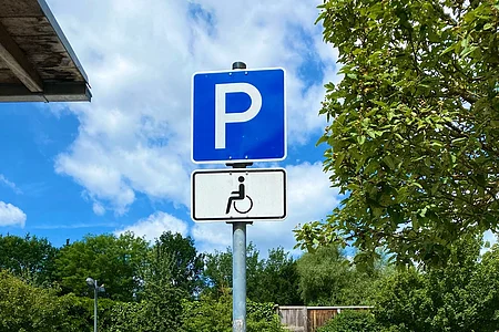 Parkplatzschild mit Rollstuhlsymbol auf einem Parkplatz mit mehreren geparkten Autos und Bäumen im Hintergrund.