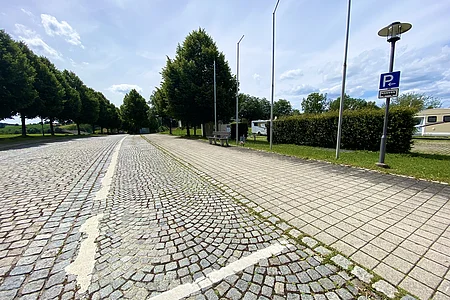 Parkplatz mit Kopfsteinpflaster und Gehweg mit Bänken, Bäumen und Wohnmobilen im Hintergrund bei Tageslicht