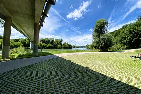Unter einer Brücke mit Pfeilern ein gepflasterter Weg, im Hintergrund Fluss und bewaldete Ufer unter blauem Himmel mit Wolken.