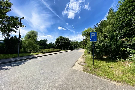 Straße mit Parkhinweisschild links, Bäume rechts und links, blauer Himmel mit Wolken.