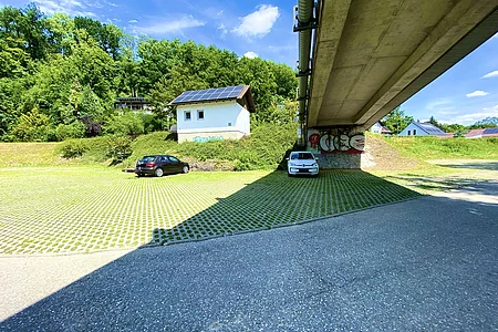 Parkplatz mit zwei Autos unter einer Brücke, im Hintergrund ein Haus mit Solarpanel und Bäumen bei sonnigem Himmel