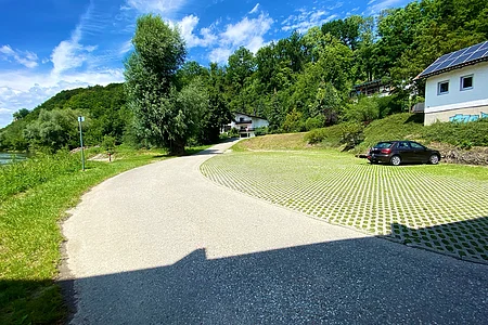 Asphaltstraße mit Parkplatz aus Rasengittersteinen, einem schwarzen Auto und Häusern vor bewaldetem Hügel unter blauem Himmel