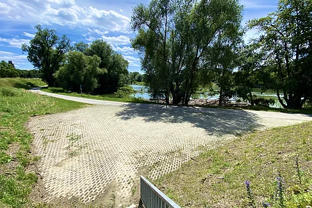 Parkplatz mit Gittersteinen neben einem Fluss, umgeben von Bäumen und Wiese unter blauem Himmel mit Wolken