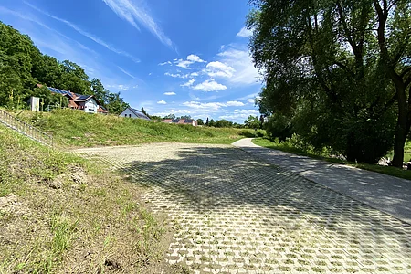 Weg mit gepflastertem Parkplatz links und Bäumen rechts unter blauem Himmel mit Wolken und Häusern im Hintergrund.