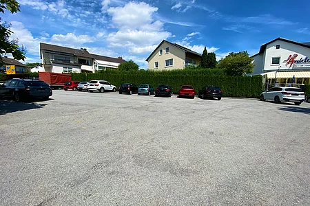 Parkplatz mit mehreren geparkten Autos vor Wohnhäusern und einem Café bei blauem Himmel und Wolken