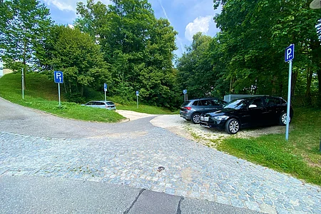 Parkplätze auf geschottertem Boden am Waldrand mit mehreren blauen Parkplatzschildern und geparkten Autos.