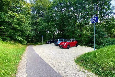 Parkplatz mit drei geparkten Autos neben einem Waldweg und Bäumen im Hintergrund