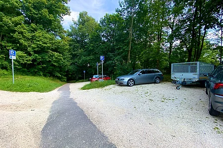 Parkplatz mit mehreren Autos und einem Anhänger neben einem Weg, umgeben von Bäumen und Grünflächen.