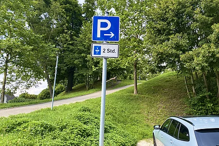 Blauer Parkplatzschild mit Pfeil nach rechts und Zusatzschild Parkscheibe 2 Stunden, umgeben von Bäumen und Wiese, rechts geparktes Auto.