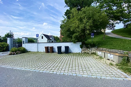 Parkplatz mit vier Mülltonnen vor weißer Mauer, zwei Schilder für Behindertenparkplätze, Bäume und Haus im Hintergrund