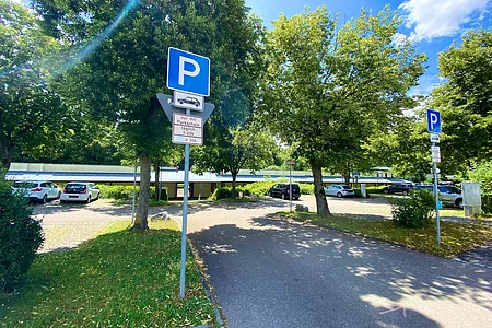 Parkplatz mit Bäumen, Autos und Parkhinweisschildern bei sonnigem Wetter und blauem Himmel.