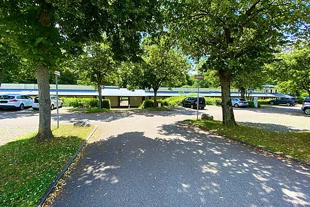 Parkplatz mit mehreren geparkten Autos und Bäumen, im Hintergrund ein langes Gebäude mit Flachdach.