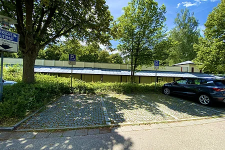 Parkplatz mit zwei Autos, Bäumen und Parkscheinautomat bei sonnigem Wetter.