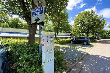 Parkautomat mit Schild „Parkscheinautomat Hier Parkschein lösen“ neben Parkplätzen und Bäumen an sonnigem Tag