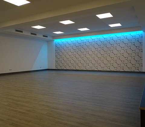 Leerer Raum mit Holzfußboden, beleuchteter Wand mit kreisförmigem Muster und Sitzkissen auf der rechten Seite.