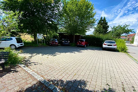 Parkplatz mit fünf geparkten Autos, Bäumen und Sträuchern bei sonnigem Wetter unter blauem Himmel