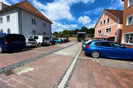 Parkplatz mit mehreren geparkten Autos vor zwei Gebäuden unter blauem Himmel mit Wolken