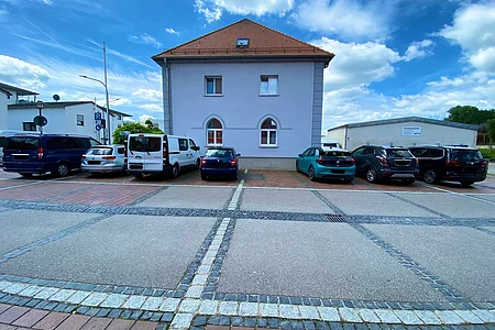 Parkplatz mit mehreren geparkten Autos vor einem zweistöckigen Haus mit rotem Dach und blauem Himmel.